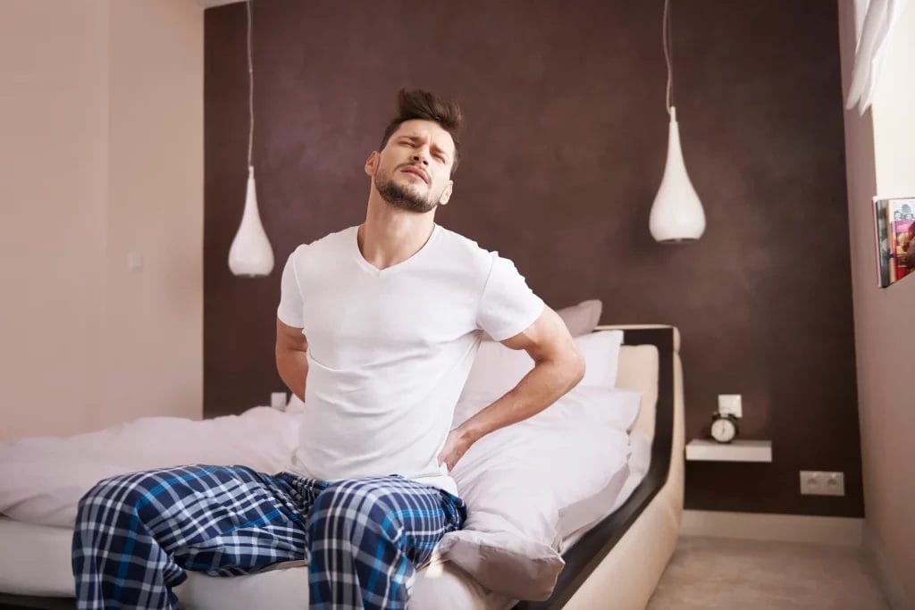 Homme assis sur un lit, en pyjama, se tenant le bas du dos avec une expression de douleur.