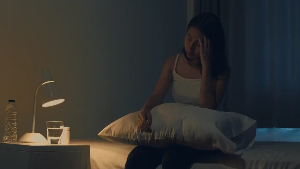 Femme assise sur un lit dans une chambre sombre, tenant un oreiller contre elle et se frottant la tempe, semblant souffrir d'insomnie ou de maux de tête, avec une lampe allumée et un verre d'eau sur la table de nuit.