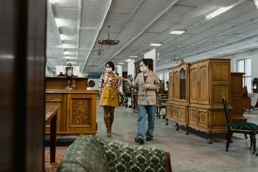  Deux femmes portant des masques de protection discutent tout en marchant dans un grand magasin de meubles anciens, entourées de buffets et armoires en bois sculpté. 